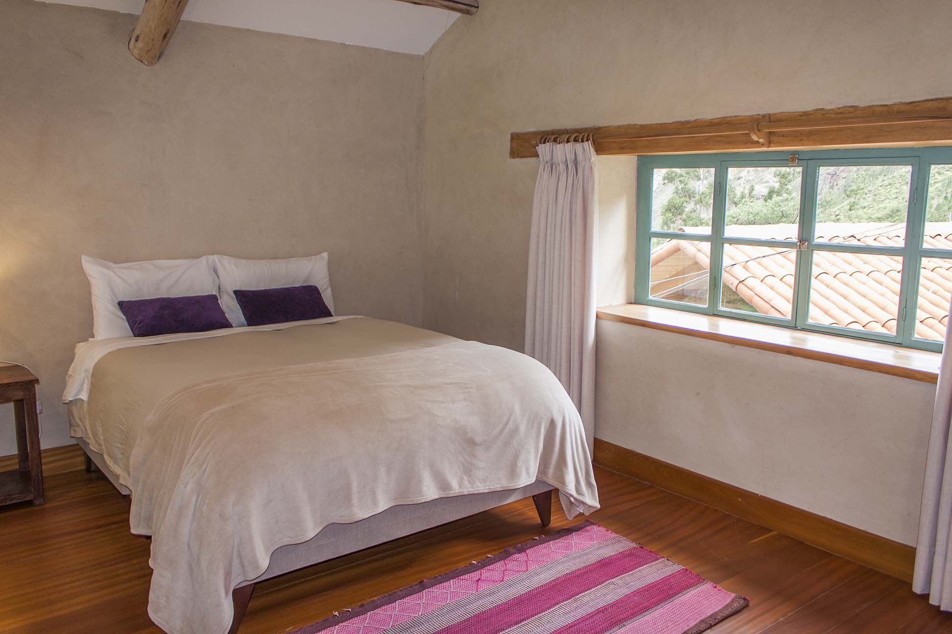A cozy bedroom with a large bed with white and purple pillows, a wooden nightstand, a window with white curtains, and a view of tile rooftops and green trees outside.