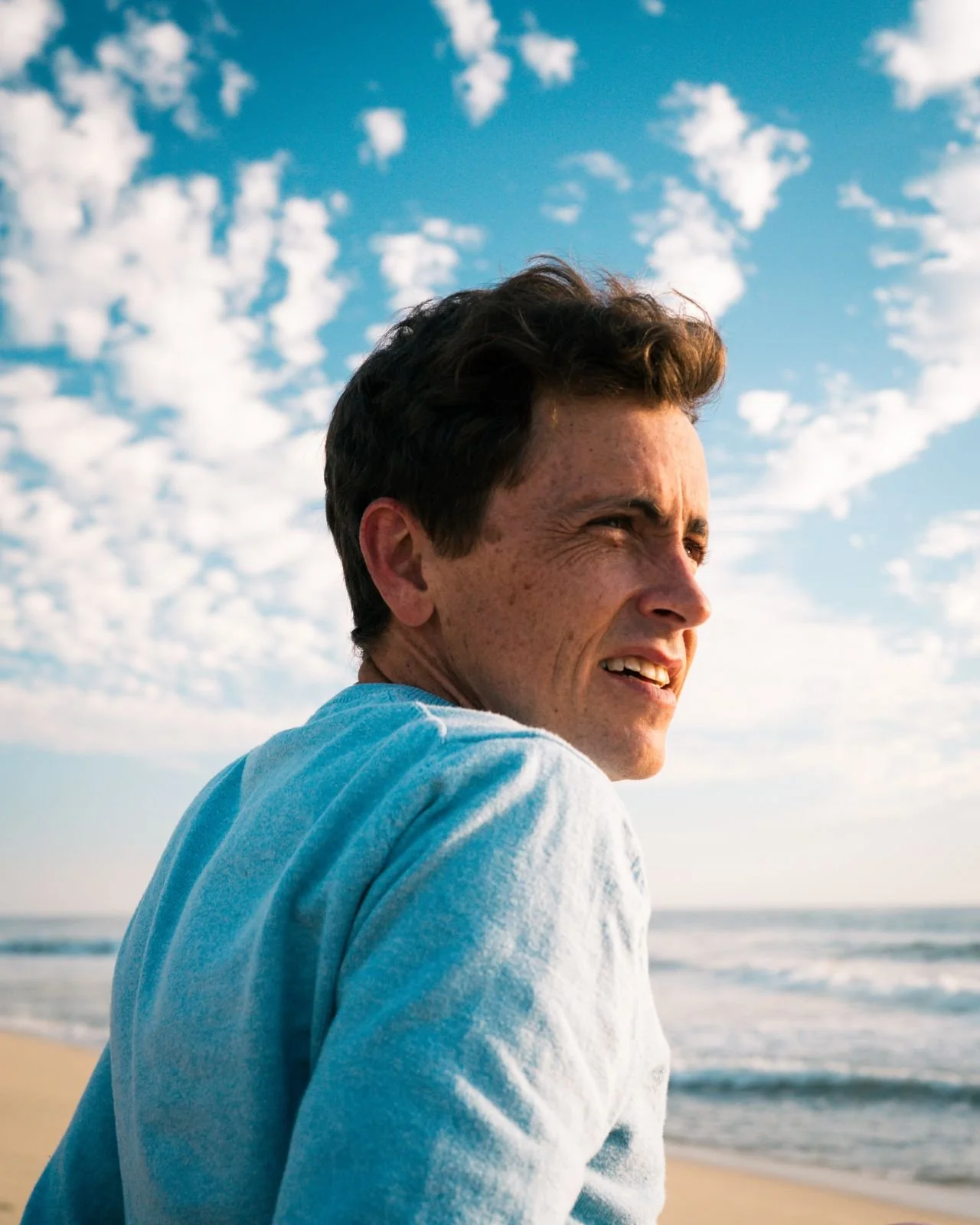 A man with dark hair looking towards the ocean on a beach, with a blue sky and scattered white clouds.