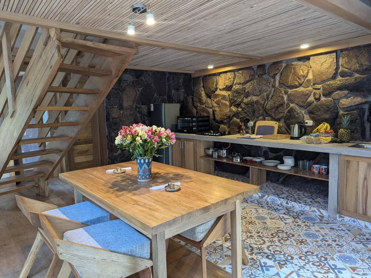 Interior of a rustic kitchen with a wooden dining table, a flower vase, a stone wall, a countertop with kitchen appliances, fruits, and patterned floor tiles.