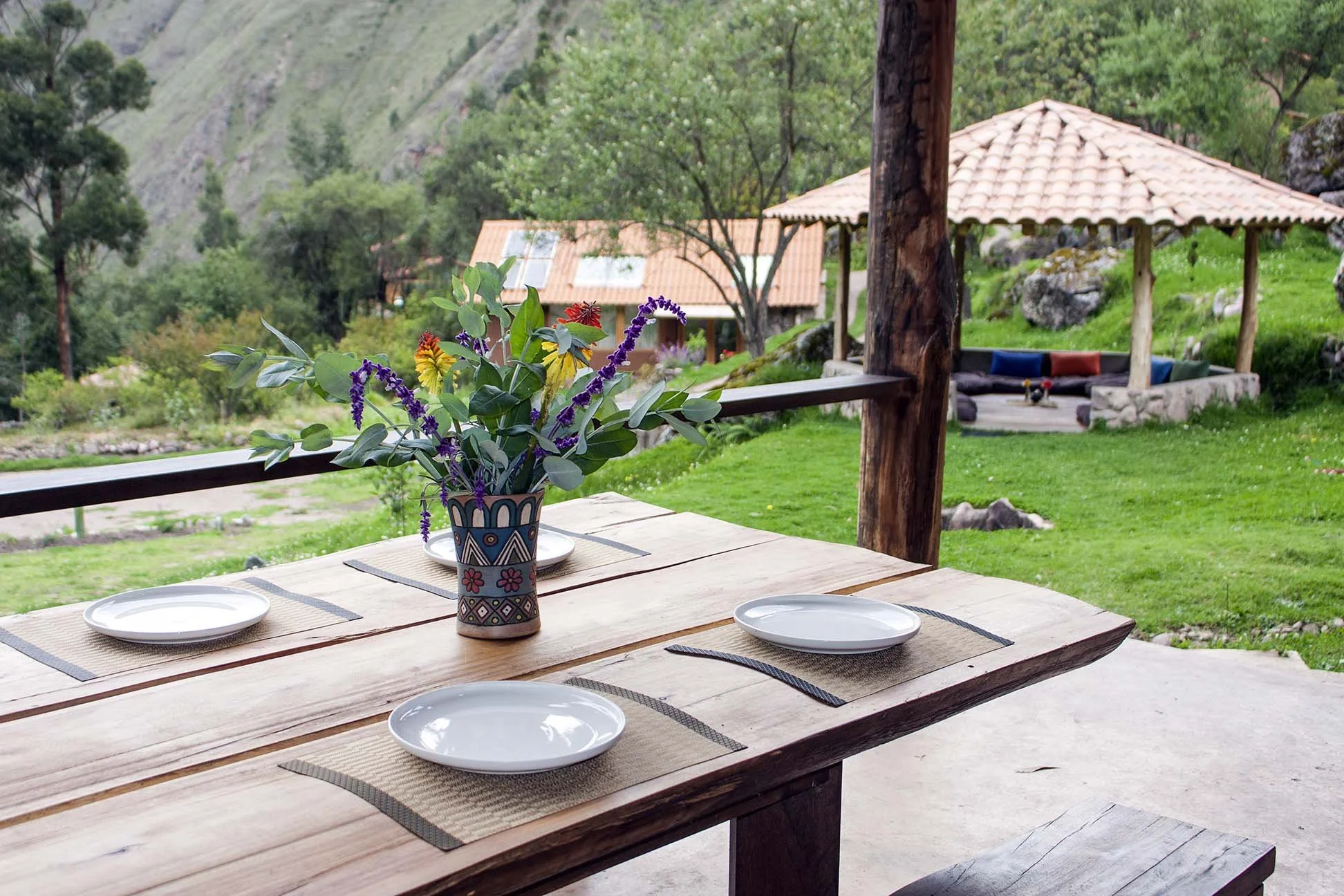 An outdoor wooden dining table set with four white plates and a colorful flower arrangement in a decorative vase, overlooking a lush green landscape with a gazebo and small building with red roofs.
