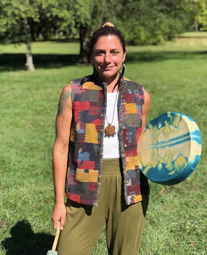 Woman standing outdoors on grass, holding a colorful round object in her left hand and a stick in her right hand, with trees in the background.