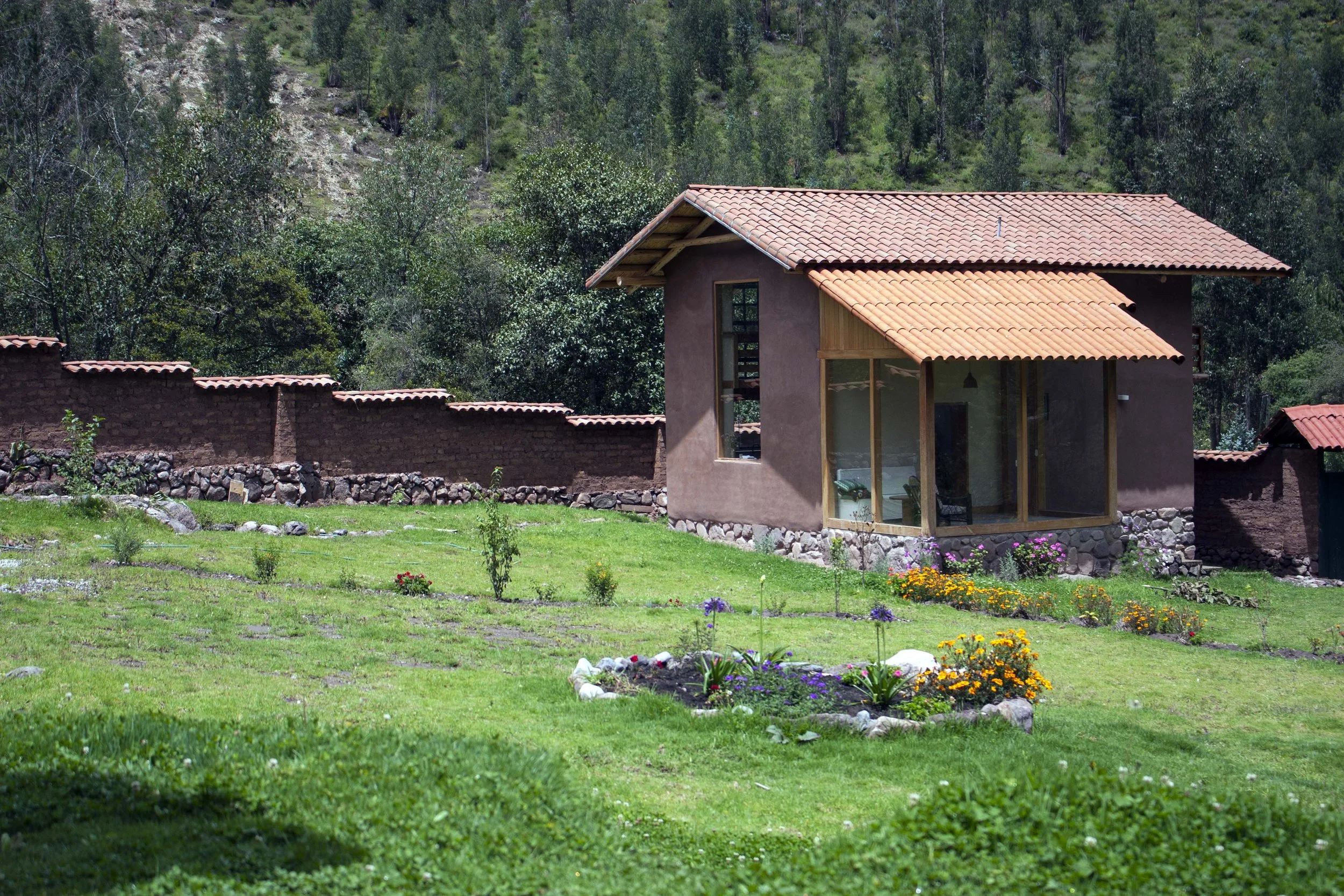 A small house with a red tiled roof and large windows is situated on a grassy lawn with flower beds. There is a stone and brick fence behind the house, and green trees and hills are in the background.