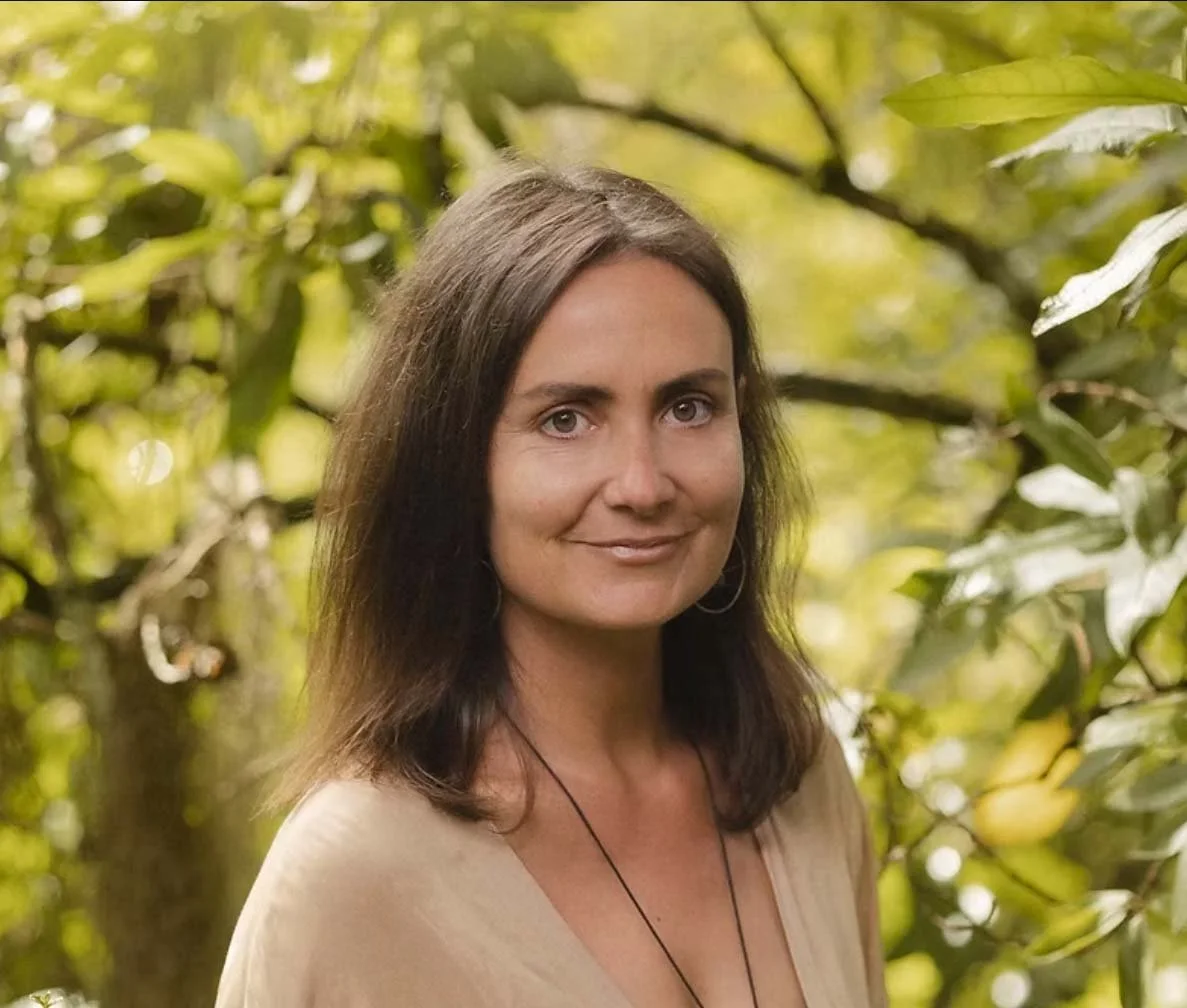 A woman with shoulder-length dark hair smiling outdoors among green trees and leaves.