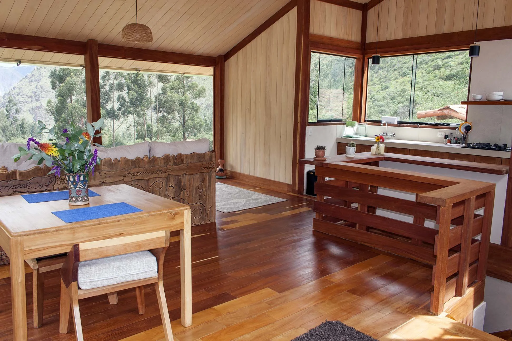 Open-concept living room and kitchen with large corner windows showing green trees and mountains outside. The space has wood-paneled walls and ceiling, a wooden dining table with a chair, a sofa with decorative pillows, potted plants on the kitchen counter, and a wooden island or breakfast bar.