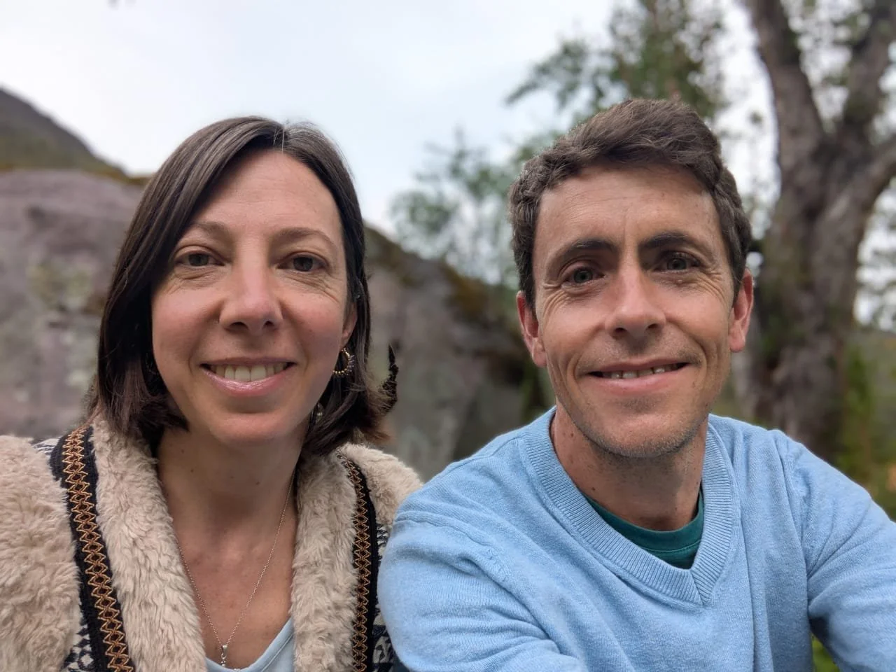 A woman and a man taking a selfie outdoors with trees and rocks in the background.