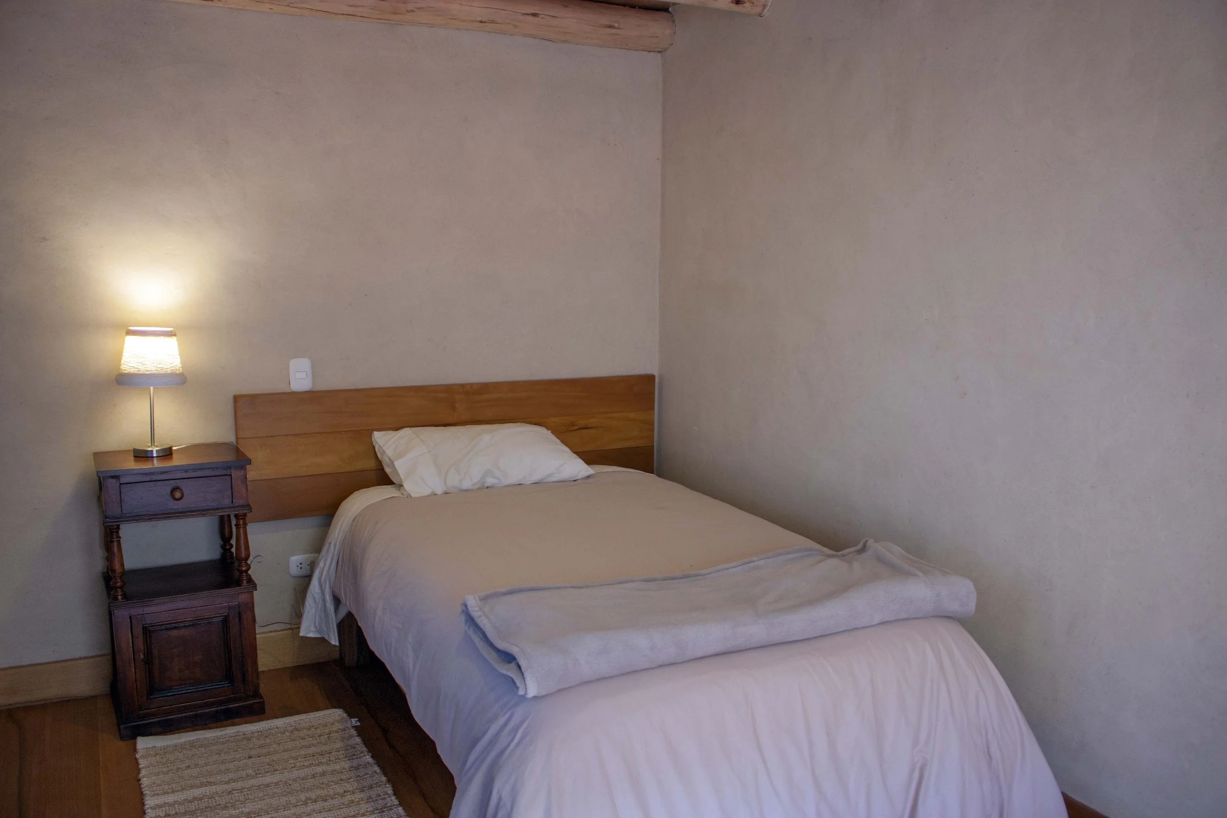 A cozy bedroom with a neatly made bed, a pillow, and a blanket at the foot. Beside the bed is a wooden nightstand with a small lamp turned on. The room has beige walls, a wooden floor, and a small rug at the foot of the bed.