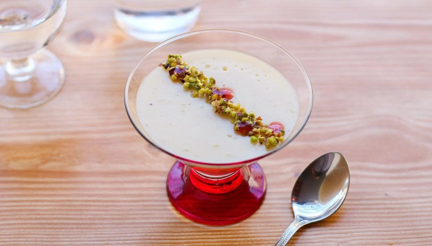 Un verre à cocktail rouge contenant une crème ou un dessert blanc, garni de pistaches hachées et d'une sauce rouge, placé sur une table en bois clair.
