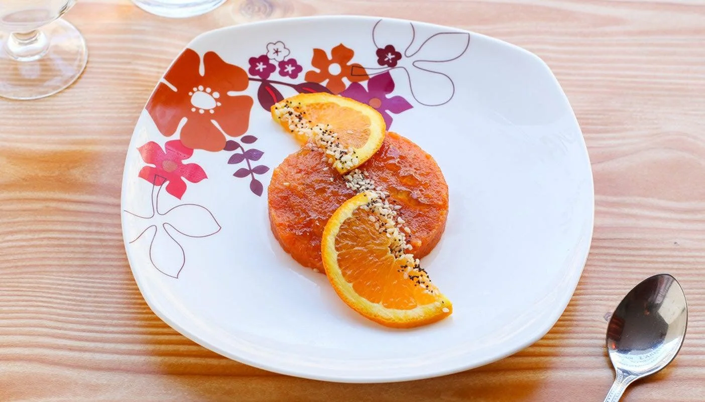 Une assiette blanche décorée de fleurs rouges, orange et violettes contient des tranches d'orange et un gâteau à l'orange, avec des graines ou des graines de sésame dessus. La table est en bois clair, avec une cuillère en métal à droite de l'assiette