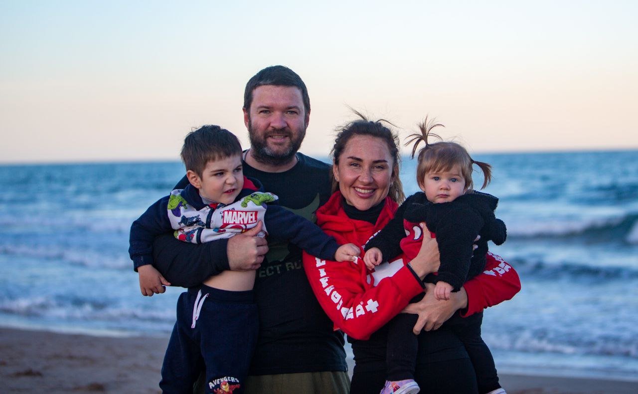 A smiling family of four standing on the beach with the ocean behind them, two children in the arms of their parents, a man and a woman, during sunset.