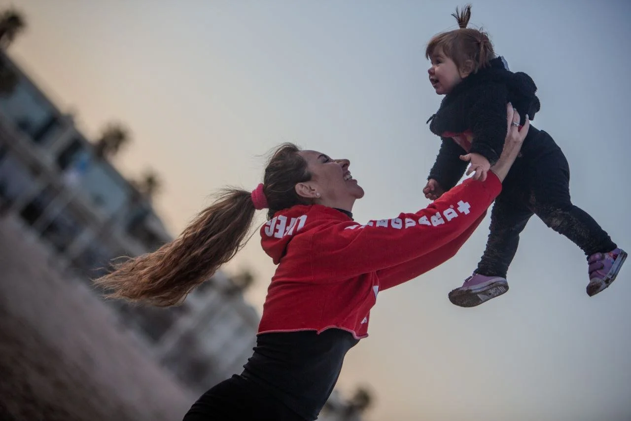 A woman with a ponytail wearing a red jacket lifts a smiling young girl in the air outdoors during dusk or evening, with buildings blurred in the background.