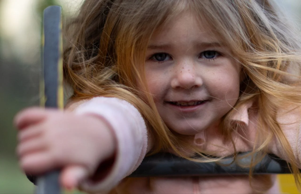 Close-up of a young girl with red hair and blue eyes smiling, reaching forward with her hand on a playground slide.