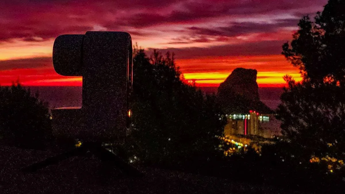 Silhouette of a traffic camera with colorful sunset sky in the background, trees on both sides, and city lights below.