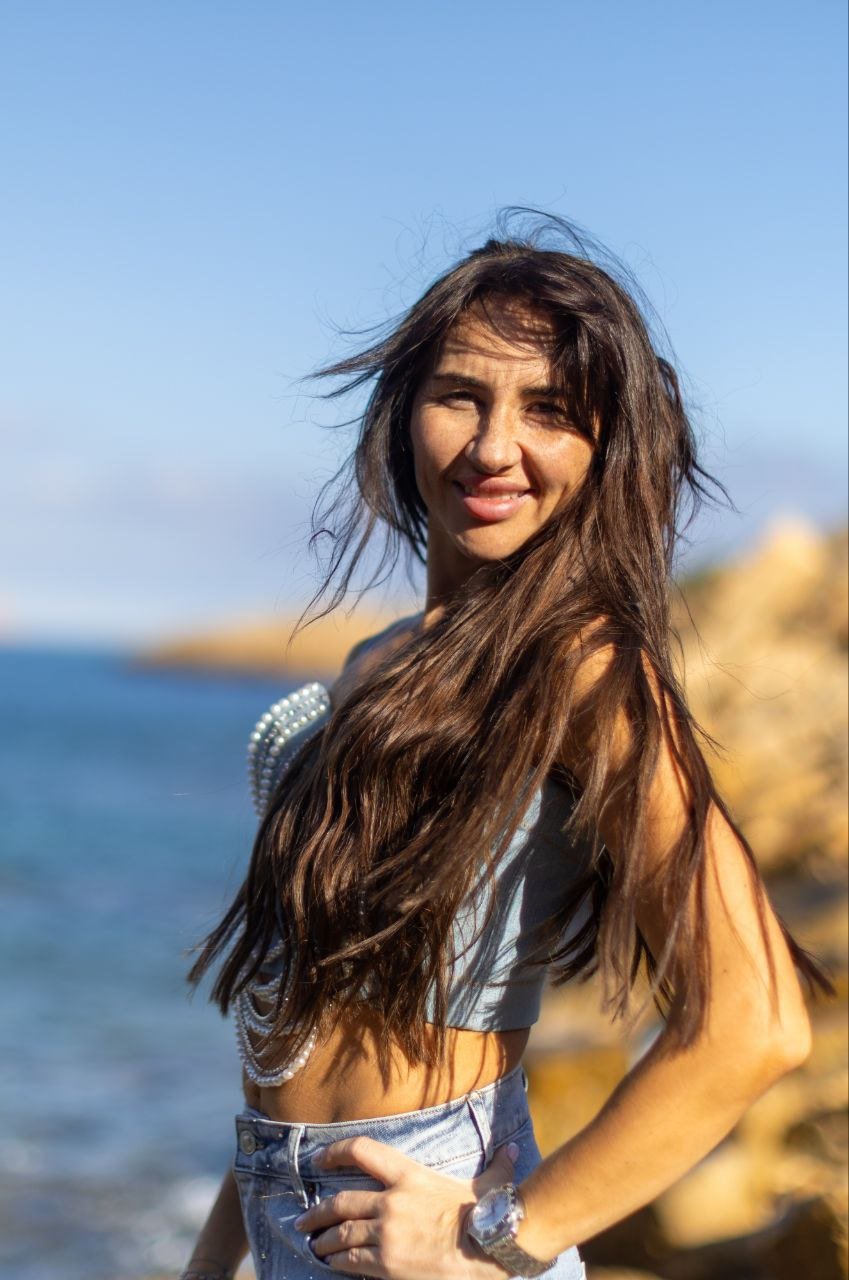 A smiling woman with long, wavy brown hair stands on a beach with rocks and water in the background. She wears a light top with pearl embellishments on the shoulder and denim shorts, looking towards the camera with a hand on her hip. The sky is blue 