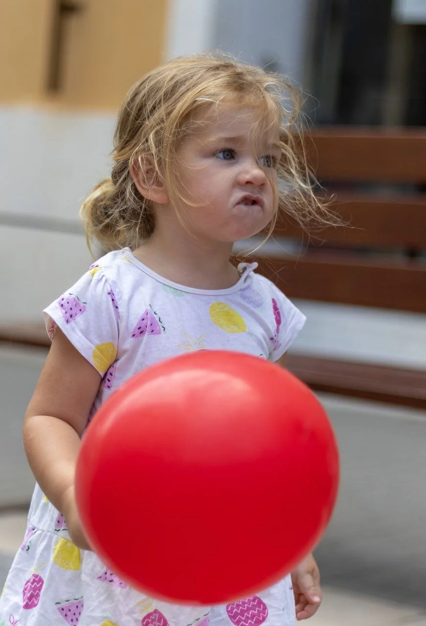 A young girl with messy blonde hair and blue eyes, making a pouty face, holding a large red balloon, wearing a white dress with colorful fruit patterns.