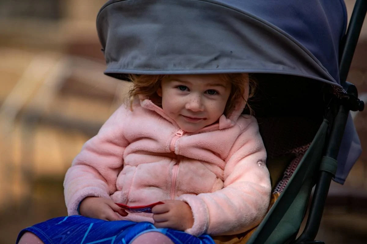 A young girl with red hair, blue eyes, and freckles sitting in a stroller, wearing a pink fleece jacket and holding a small object in her hand. She is smiling and looking at the camera.