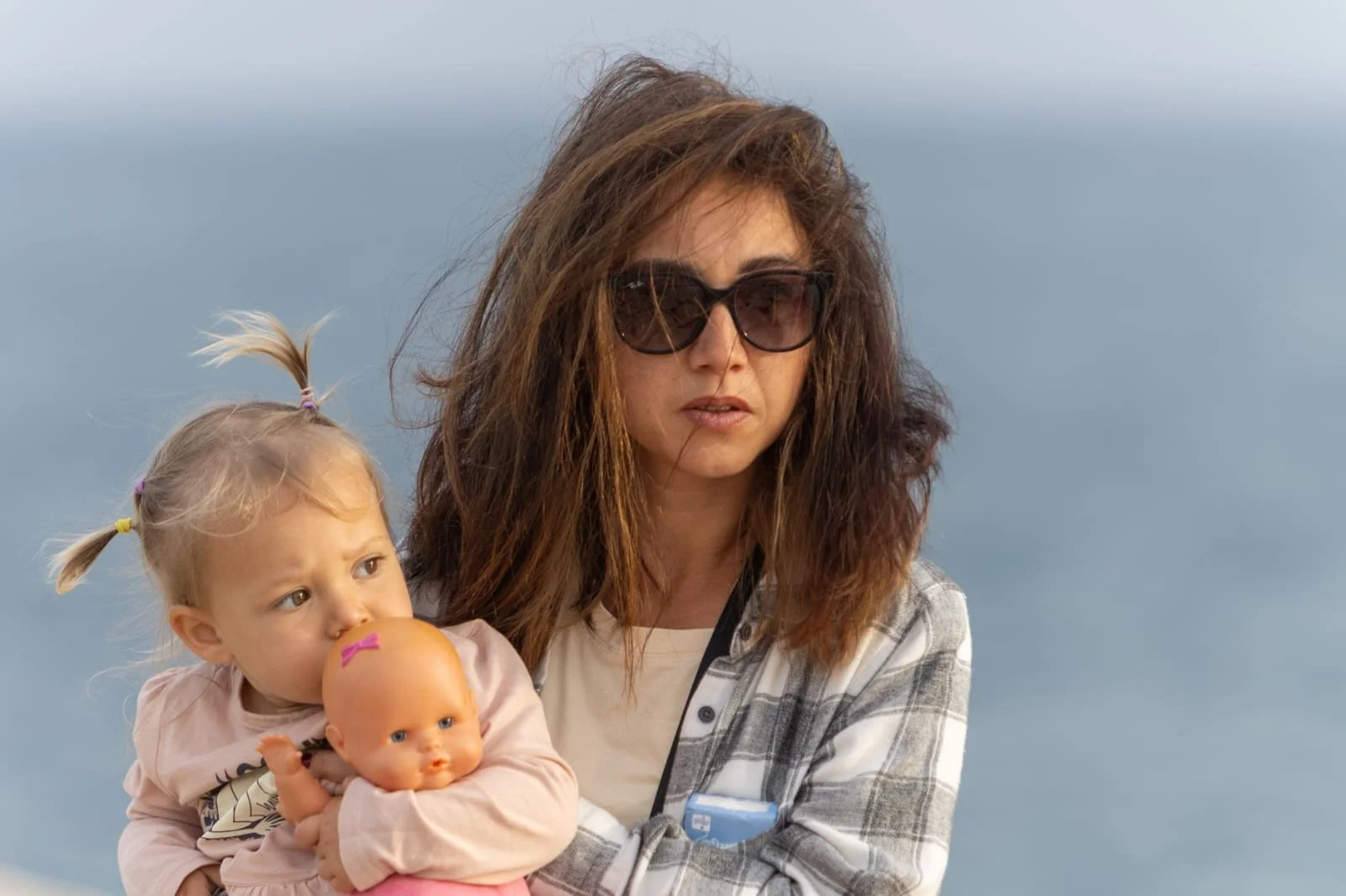 Woman with brown, wavy hair wearing sunglasses holding a young girl with pigtails and a doll, outdoors near a body of water, overcast sky.