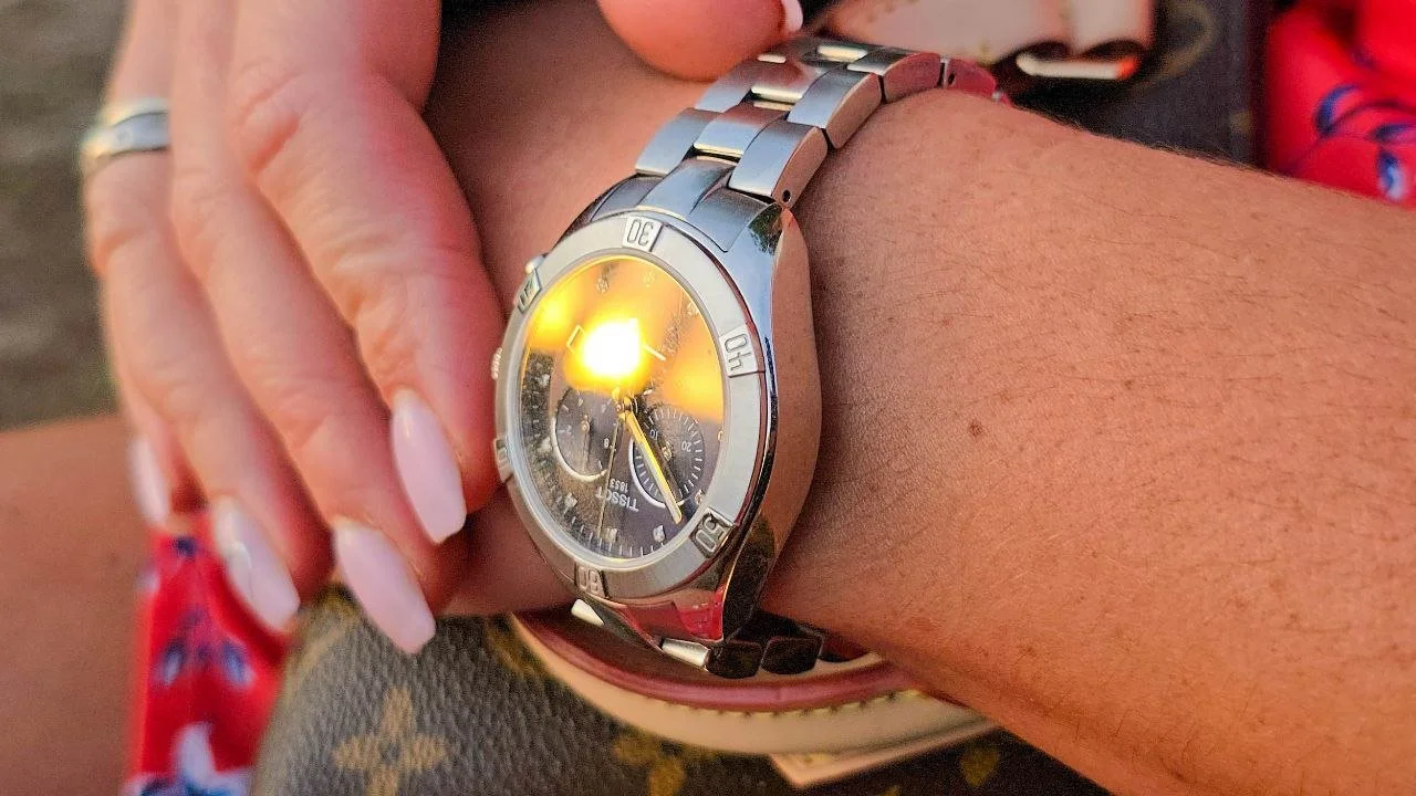 A person's left wrist wearing a silver watch with a black dial and multiple subdials, reflecting sunlight. The person has a ring on their finger and appears to be seated outdoors.