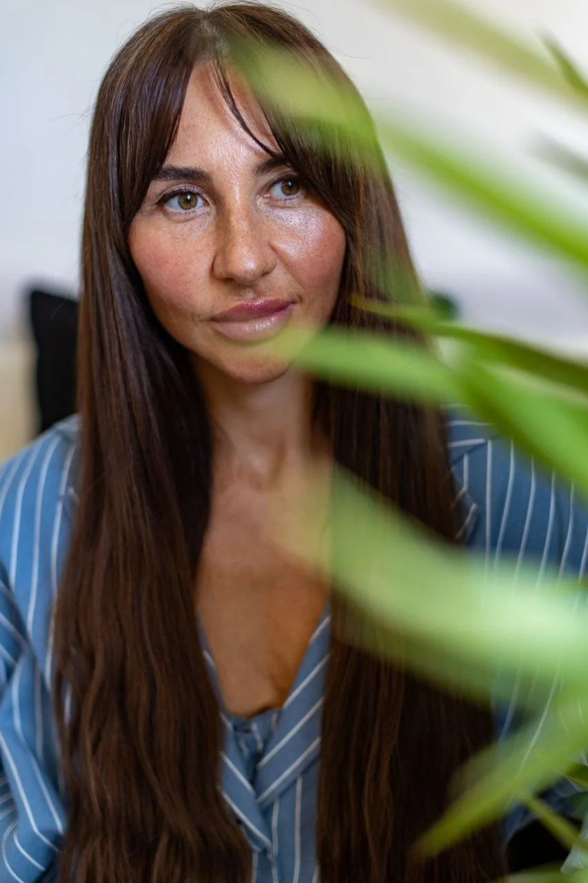 A woman with long brown hair and brown eyes looking to the side, partially obscured by green leaves, wearing a blue and white striped shirt.