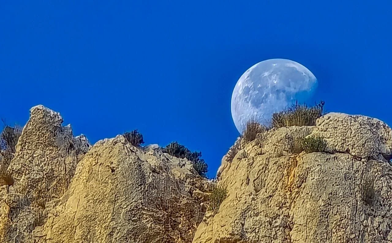 The moon in a waxing gibbous phase appears behind rocky mountain peaks, with some sparse vegetation on the rocks, all set against a clear blue sky.