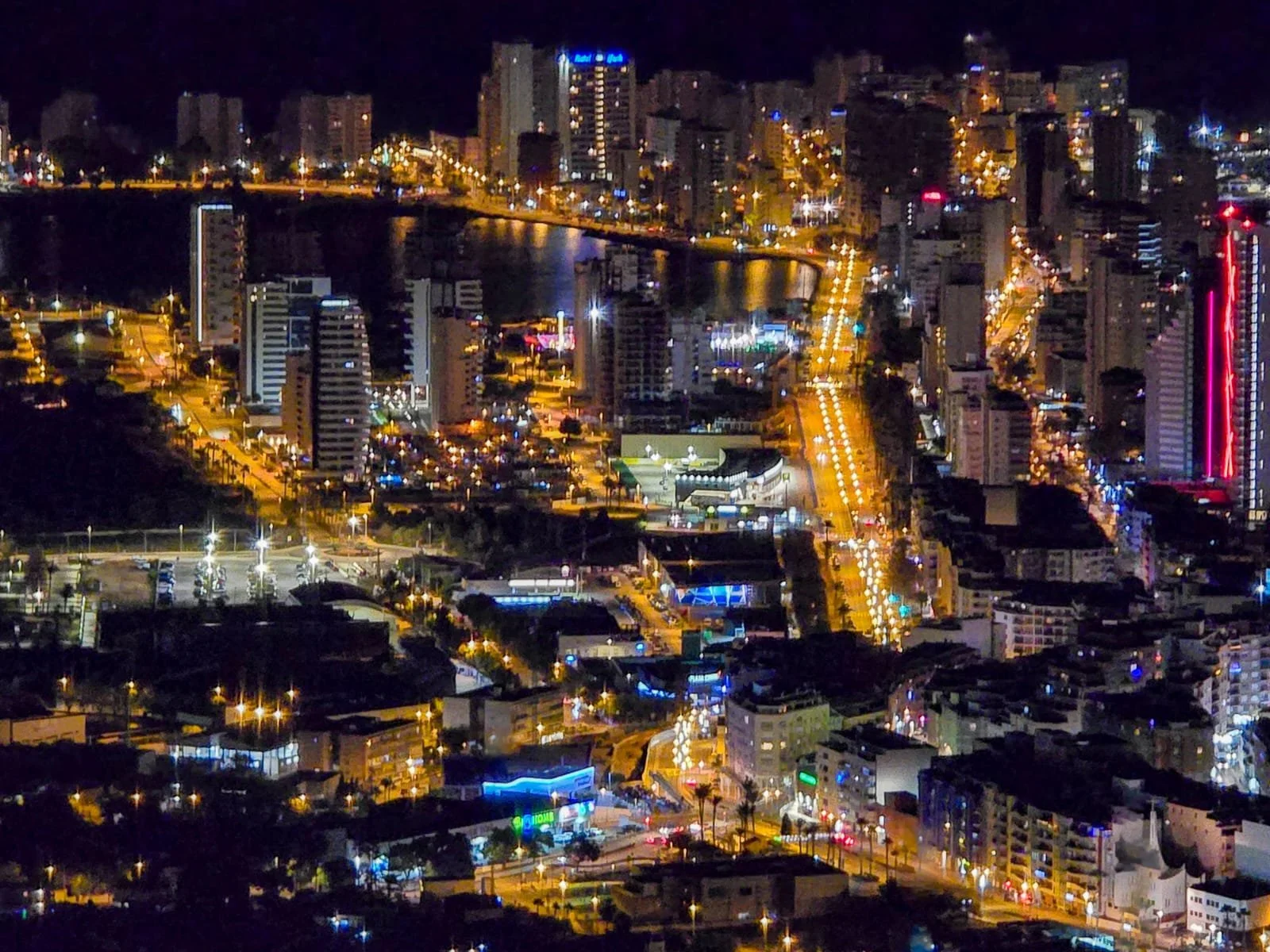 Nighttime aerial view of a city with illuminated buildings, streets, and a body of water reflecting city lights.