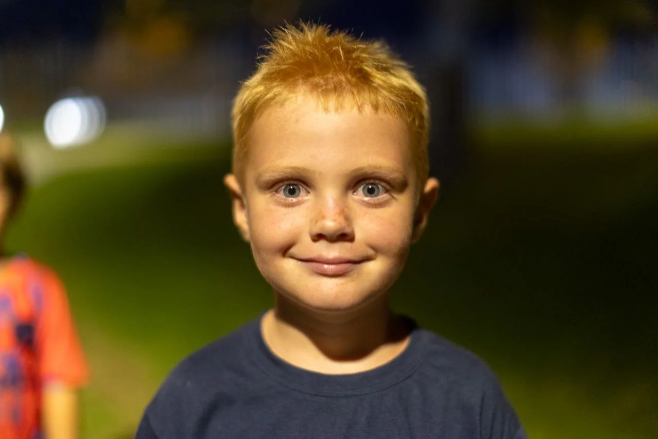 Close-up of a young boy with red hair and blue eyes smiling at night outside.