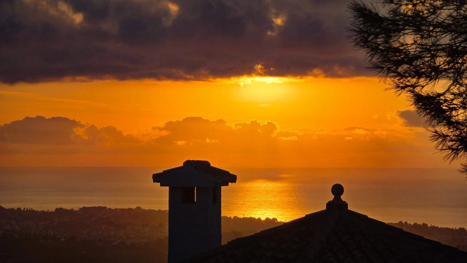 Sunset over the ocean with clouds, silhouetted rooftop and tree branches in the foreground.