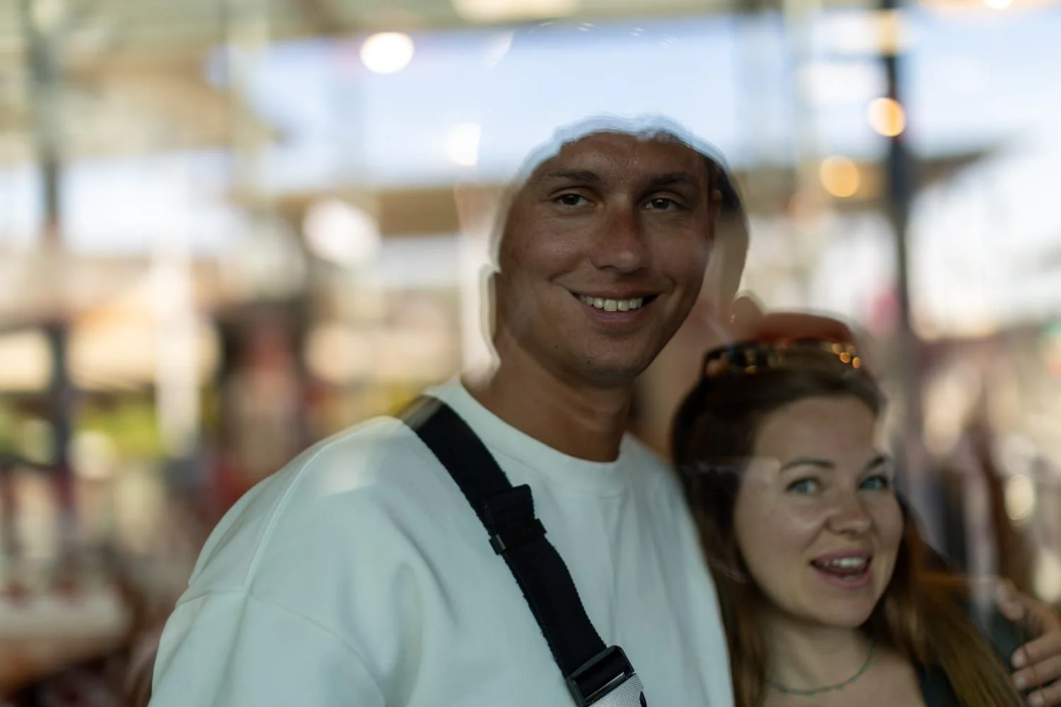 A man and woman smiling at the camera through a glass window in a brightly lit indoor setting.