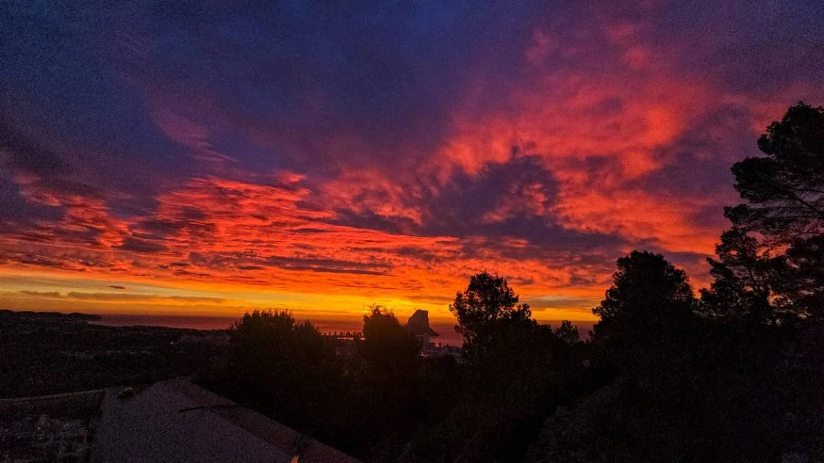 Colorful sunset sky with orange, red, and purple clouds over a silhouette of trees and distant landscape.