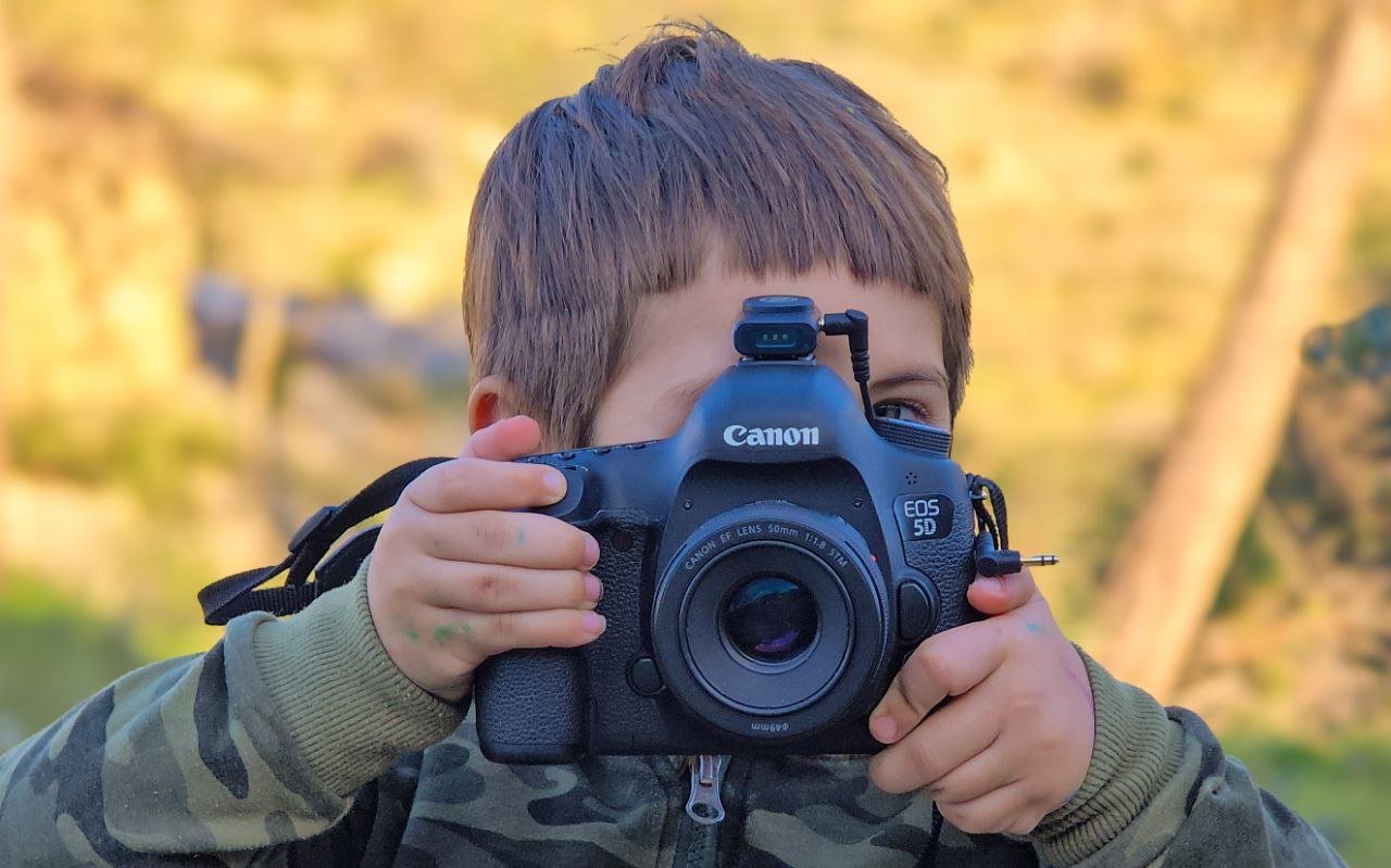 A young boy holding a Canon EOS 5D camera, aiming it towards the viewer outdoors during daytime.