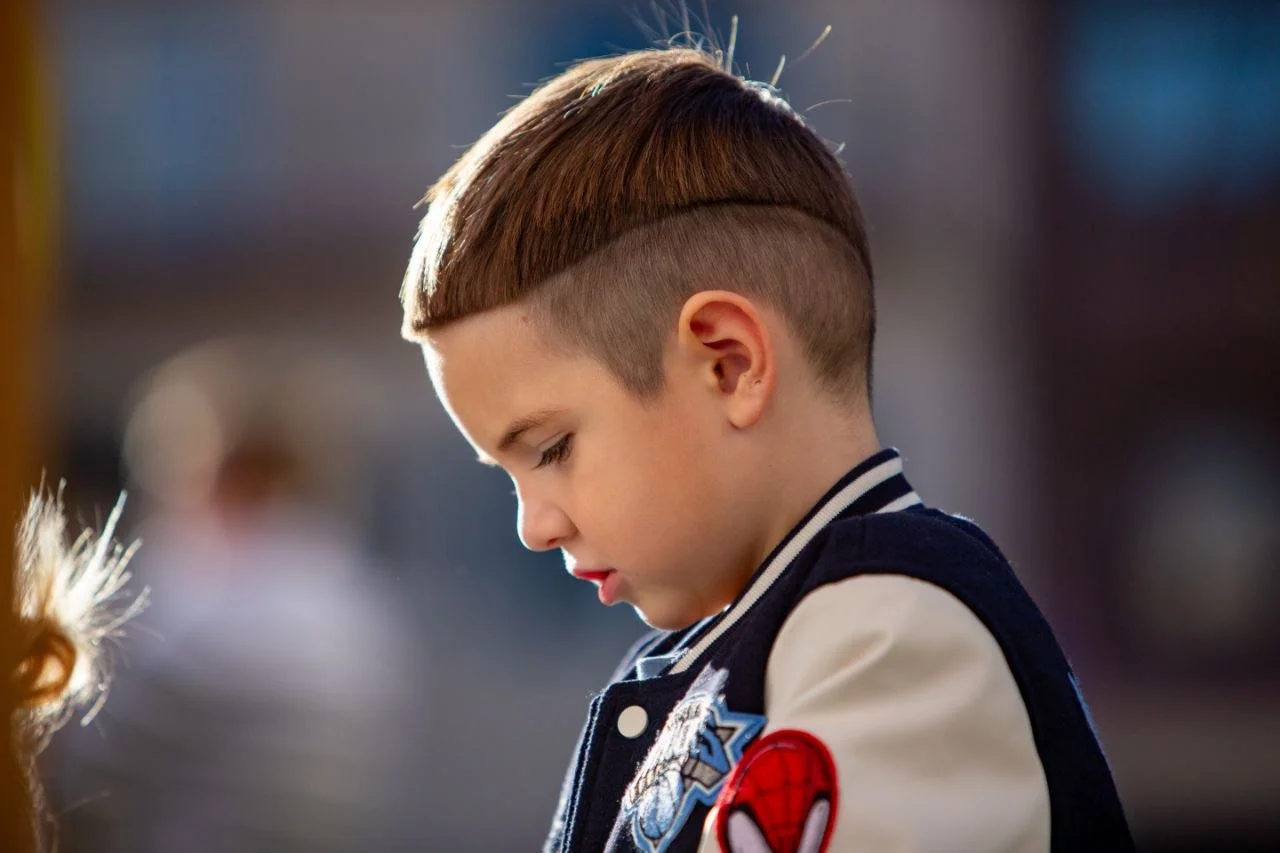 A young boy with a short haircut and a bowl cut on top, wearing a varsity jacket with a Spider-Man patch on the sleeve, looking down.