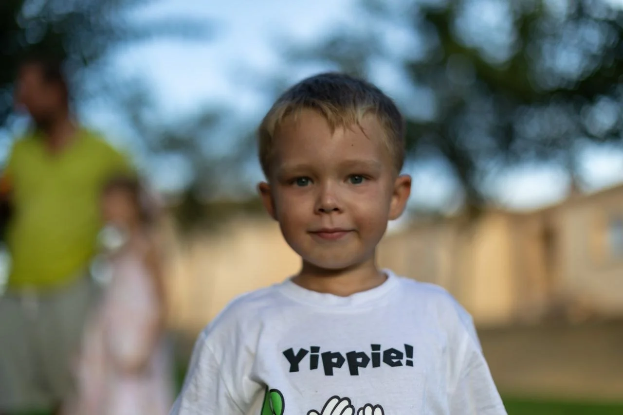 Young boy with short brown hair and blue eyes, smiling outdoors, wearing a white t-shirt with the word 'Yippie!' on it, blurred background with trees and a person in a yellow shirt.