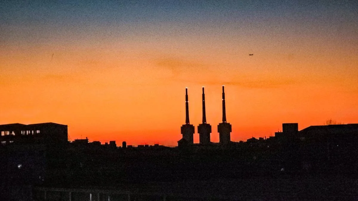 Silhouette of three industrial smokestacks against a colorful sunset sky with an airplane flying overhead.