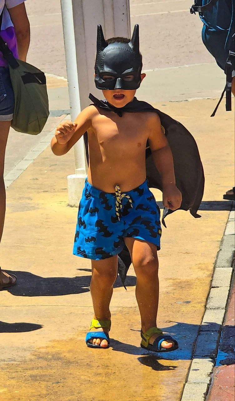 A shirtless young boy wearing a Batman mask, blue shorts with a bat pattern, and sandals, walking outdoors on a sunny day, carrying a black bag.