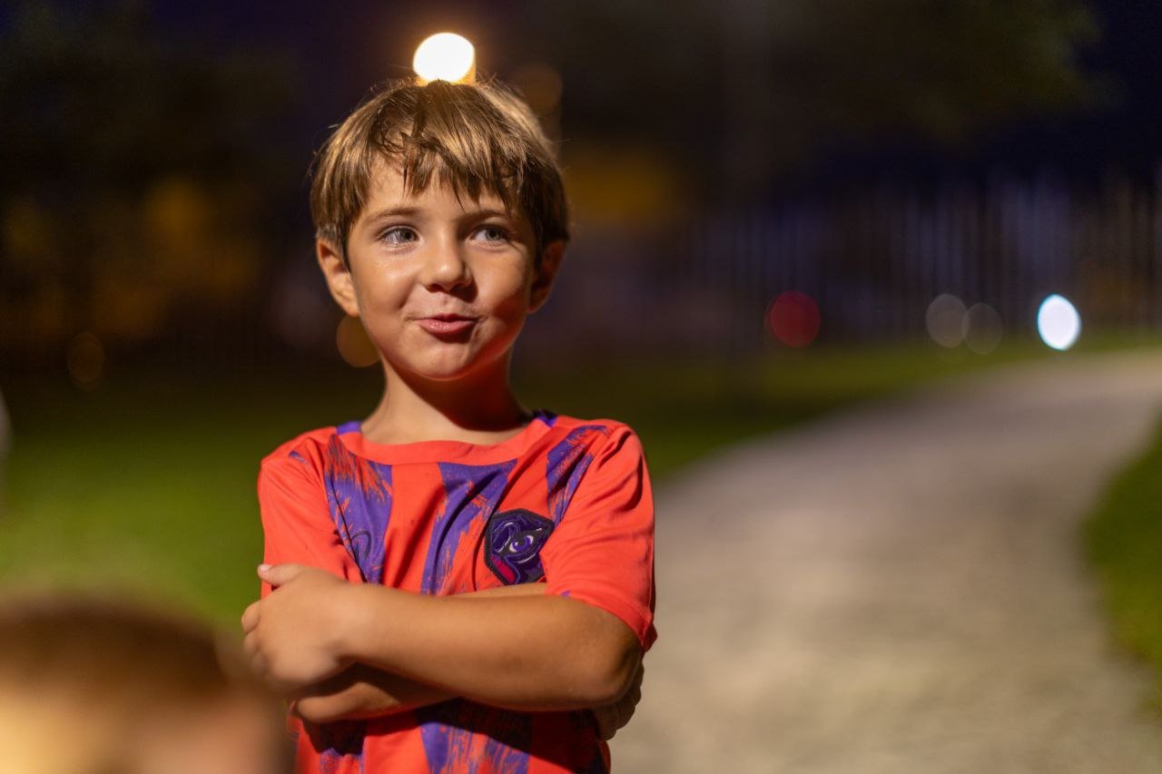A young boy with brown hair and blue eyes standing outdoors at night with arms crossed, smiling slightly, on a path with blurry lights in the background.