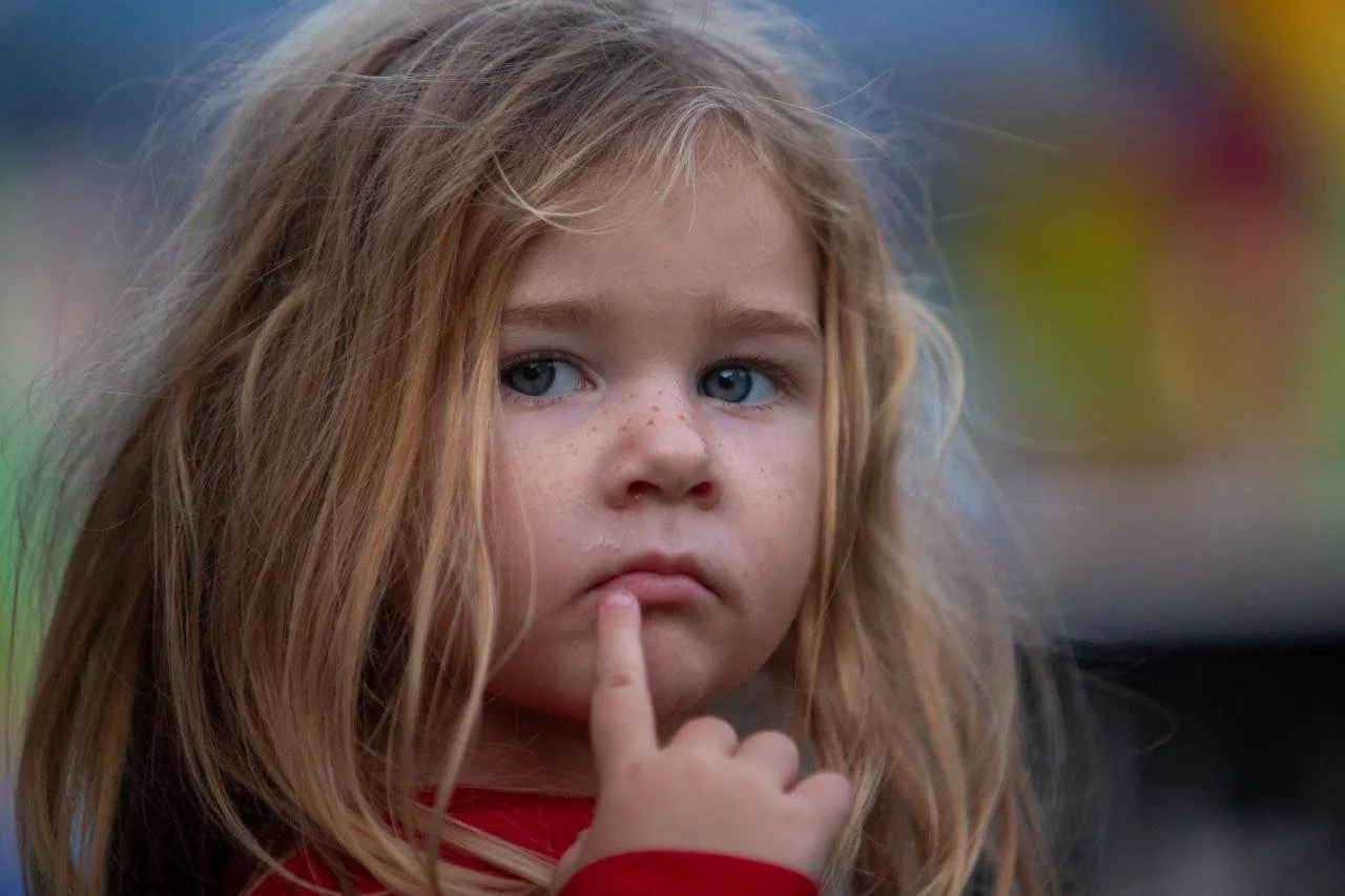 A young girl with red hair, blue eyes, and freckles touching her lips with her finger, looking thoughtful.