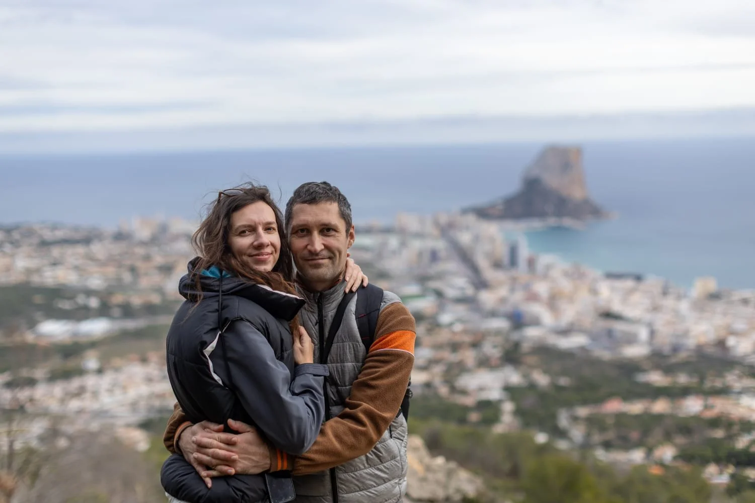 A smiling couple embracing each other with the cityscape and coastline in the background, possibly on a hill or mountain overlooking a coastal town.
