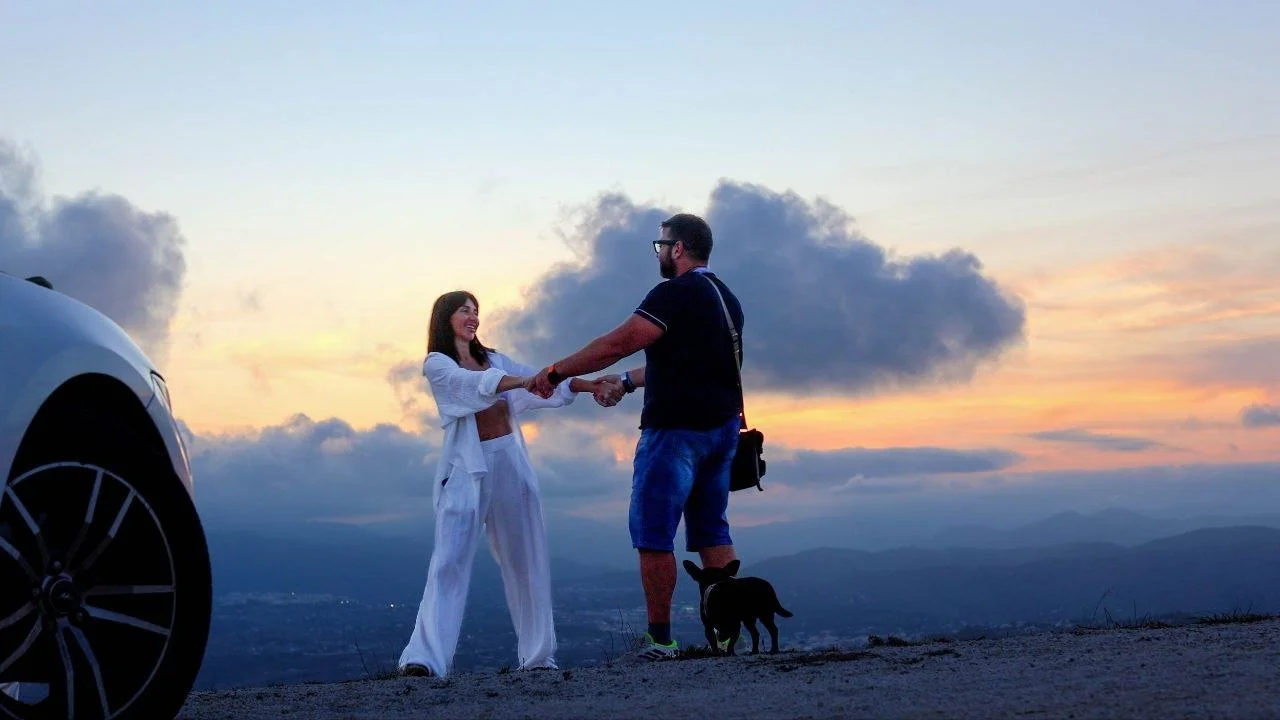 A couple exchanging a handshake outdoors at sunset, with a car on the left and a small black dog on a leash near the man's foot, rolling hills in the background.