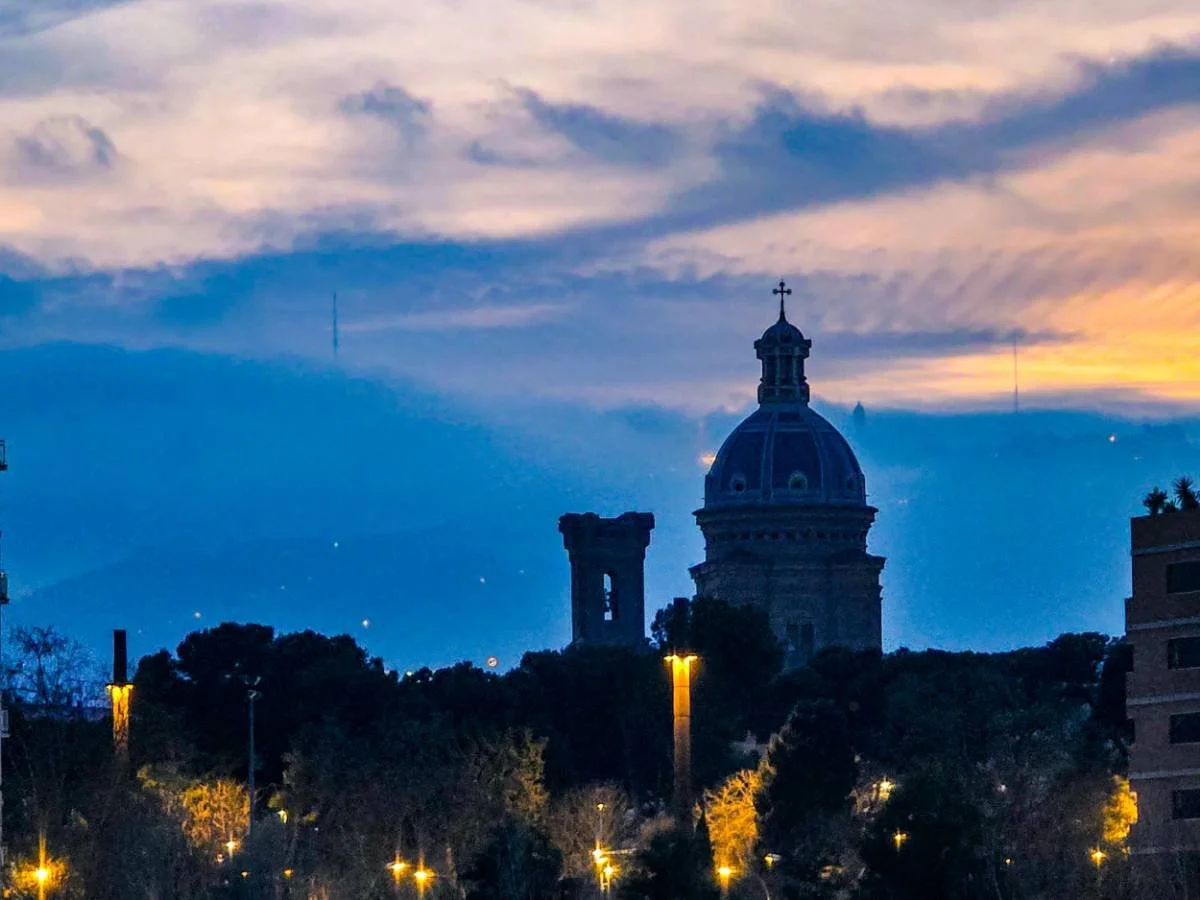 Cityscape at dusk featuring a building with a large dome and cross on top, surrounded by trees and streetlights, with a sky showing clouds and a gradient of colors from blue to orange.