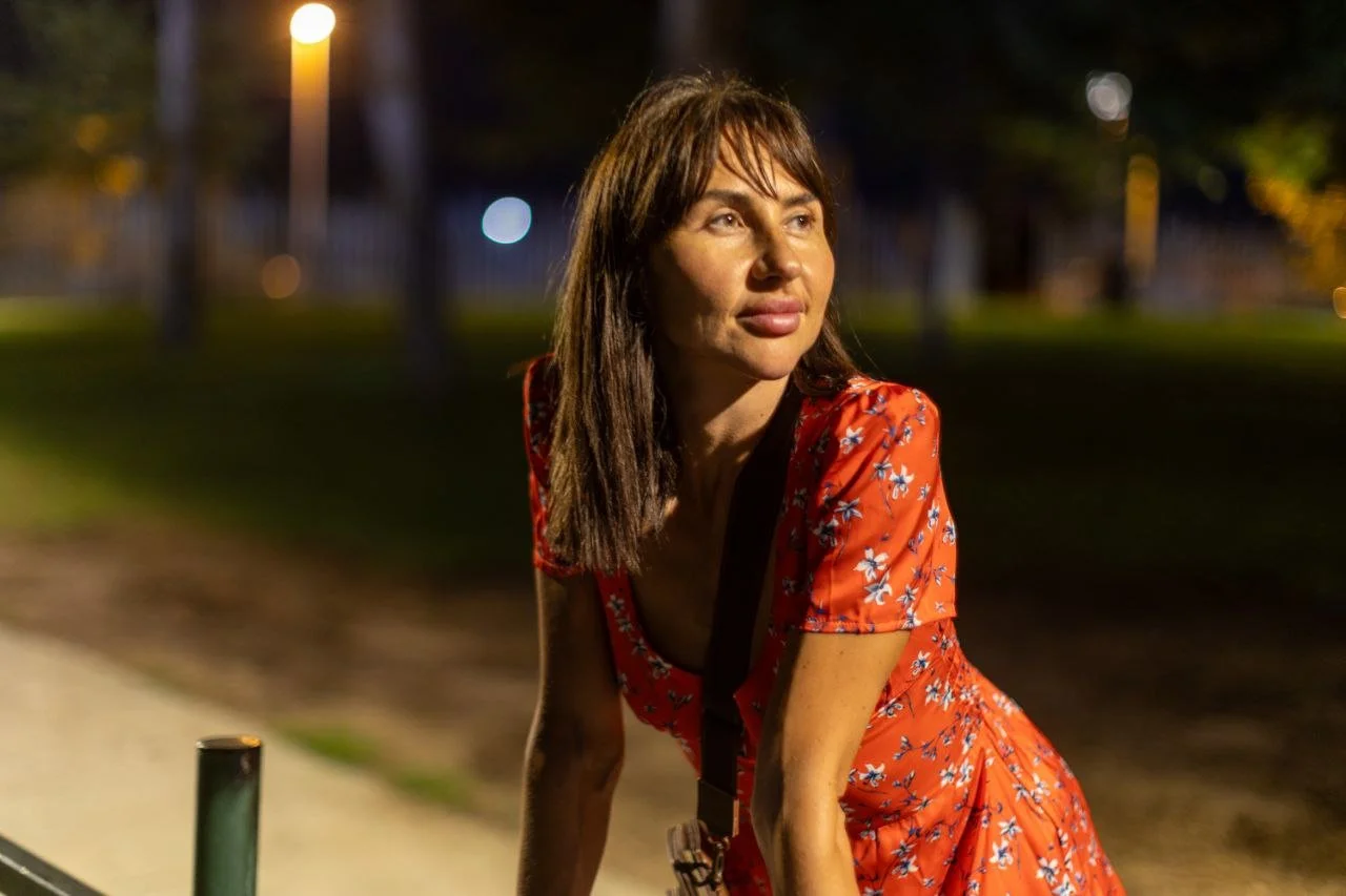A woman with shoulder-length brown hair wearing a red floral dress outdoors at night, illuminated by a streetlamp, with blurred trees and lights in the background.