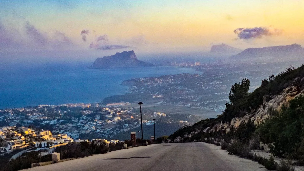 A winding road descending down a hill with houses, leading towards a bay with mountains in the background during sunset or sunrise.