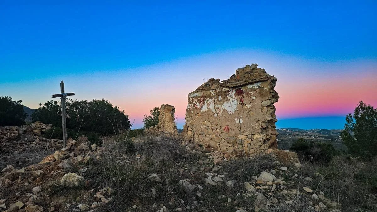 Ruins of an old stone building with a wooden cross in a rocky outdoor landscape during sunset or twilight, with colorful sky in the background.