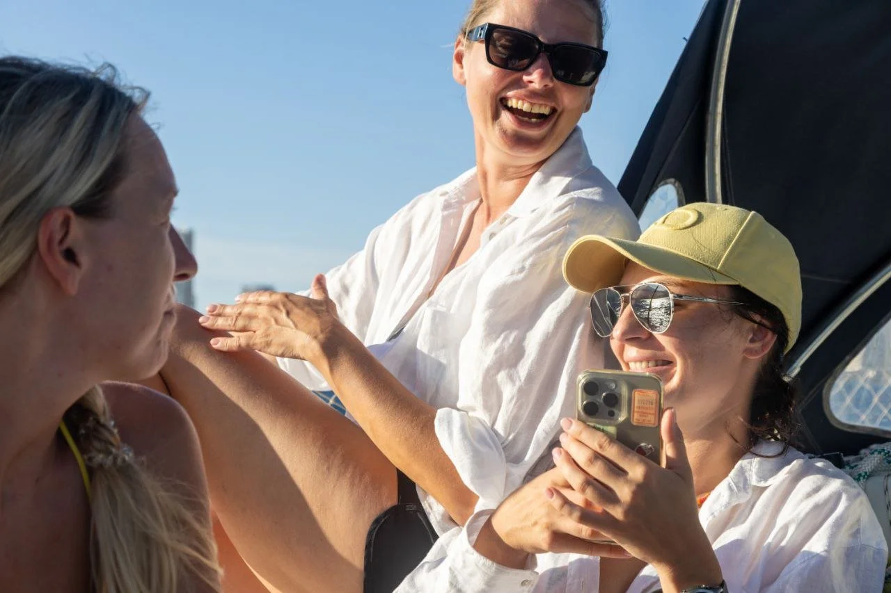Three women outdoors, one wearing sunglasses and a yellow cap, holding a smartphone, smiling, and a second woman with sunglasses, smiling, sitting next to her, and a third woman with long blonde hair, also outdoors.
