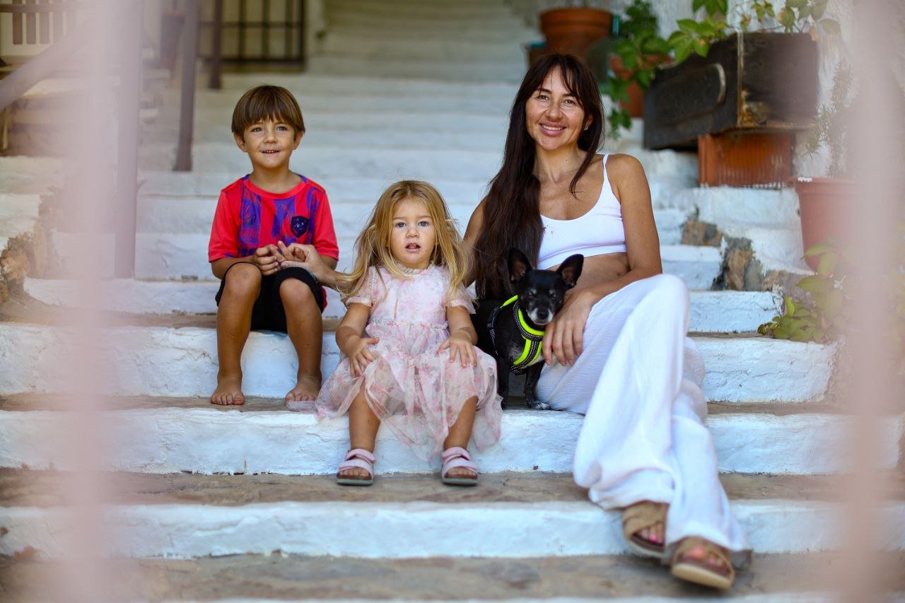 A woman sitting on white stone steps with two children, a boy and a girl, and a black dog with a harness. The boy is smiling, the girl looks surprised, and the woman is smiling. There are potted plants around the stairs.