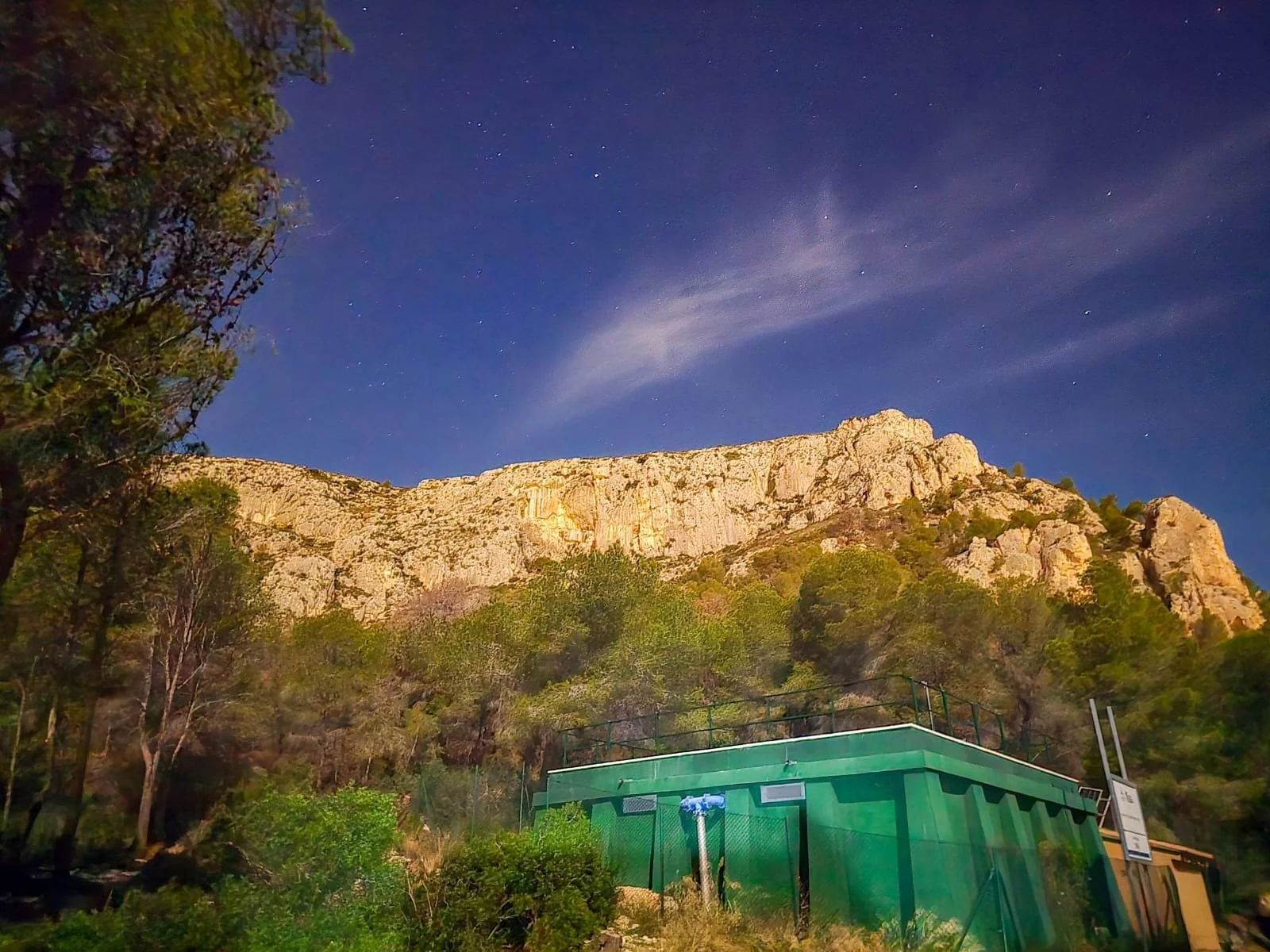 Nighttime view of rocky mountain with trees in foreground and a blue sky with stars.