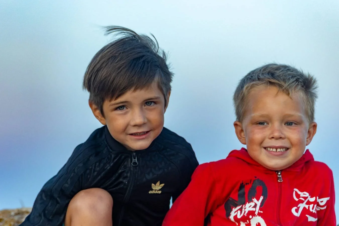 Two young boys smiling outdoors against a clear sky, one with dark hair in a black jacket and the other with light hair in a red hoodie.