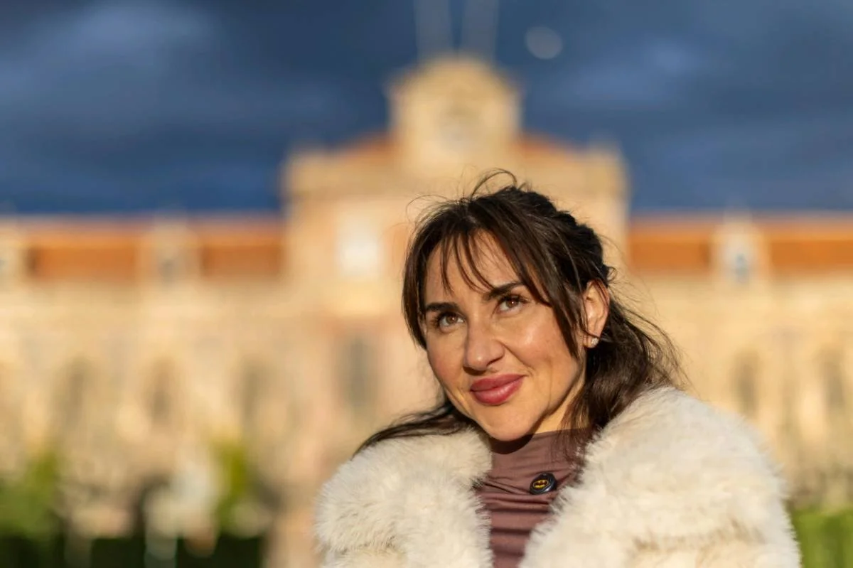A woman with dark hair and bangs, wearing a cream fur coat, smiling in front of a blurred historic building with a blue sky background.