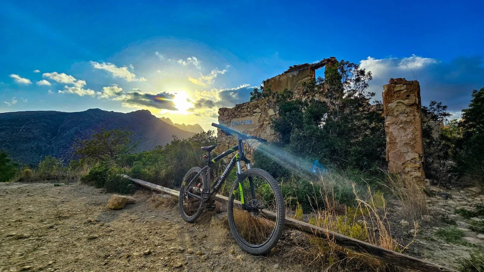 A mountain bike resting on a dirt trail near the ruins of an old stone building at sunset, with mountains and a partly cloudy sky in the background.