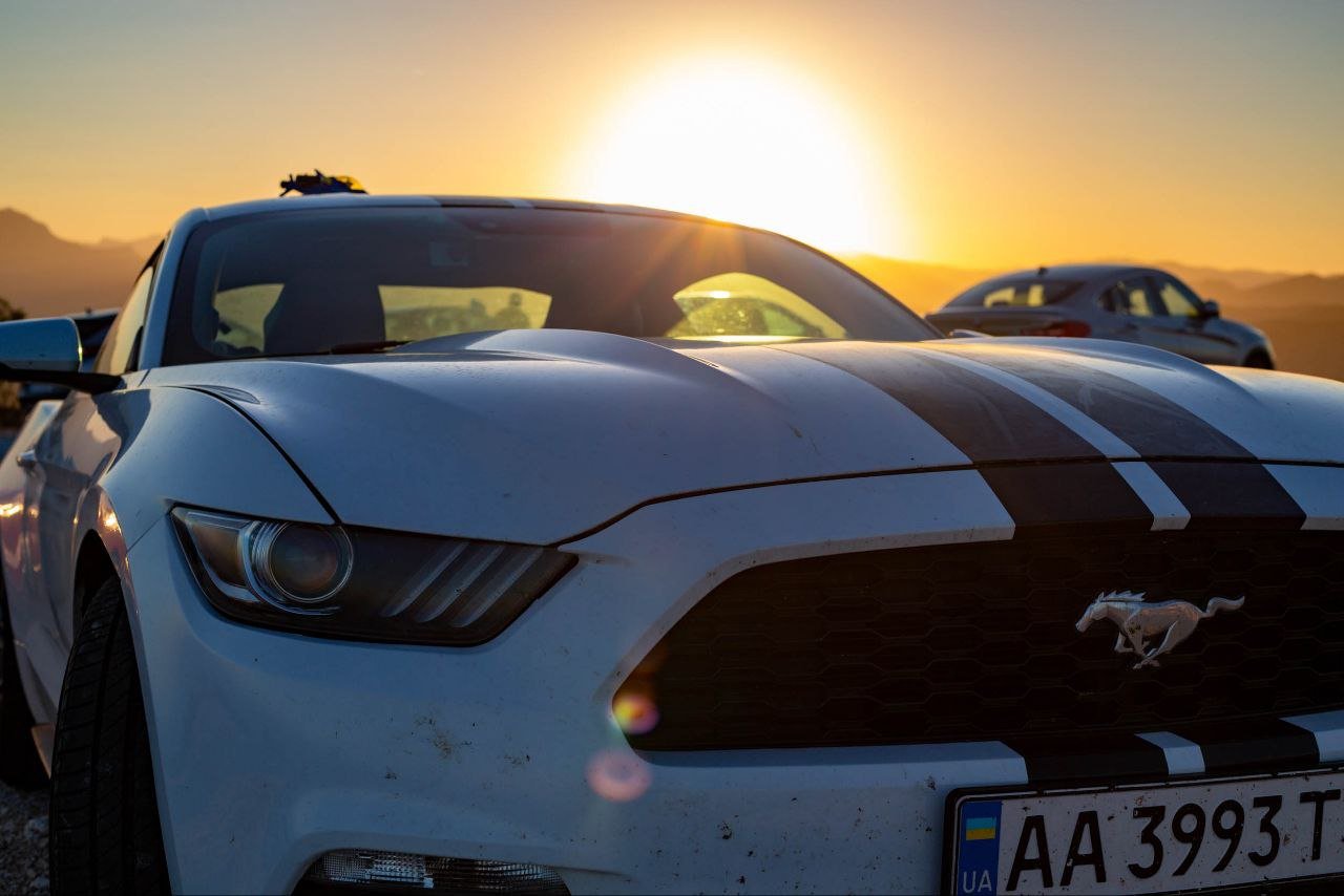 White sports car with black racing stripes parked outdoors during sunset, with other cars in the background.