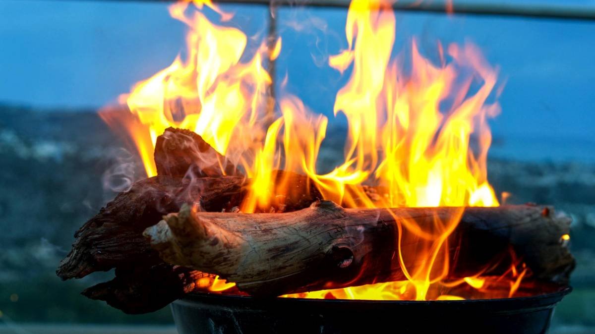 Close-up of a wood log burning with bright orange and yellow flames against a blurred ocean background.