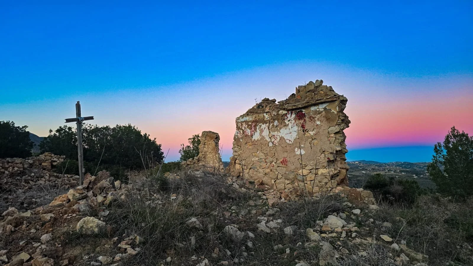 Ruins of an old stone building with a wooden cross to the left, set against a colorful sunset sky in a rural landscape with bushes and hills in the background.